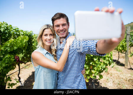 Couple heureux en tenant par selfies téléphone mobile au vignoble contre ciel bleu clair Banque D'Images