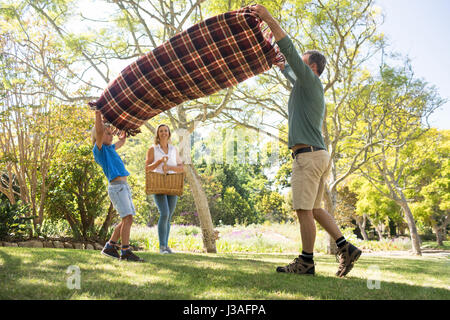 Père et fils se propager la couverture de pique-nique alors que mère carrying basket in park Banque D'Images