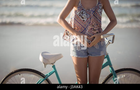 Mid section of woman making heart shape with hands standing by bicycle at beach Banque D'Images