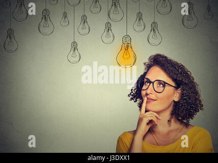 Portrait femme penser dans les verres à la lumière de l'ampoule jusqu'à l'idée au-dessus de la tête isolé sur fond de mur gris Banque D'Images