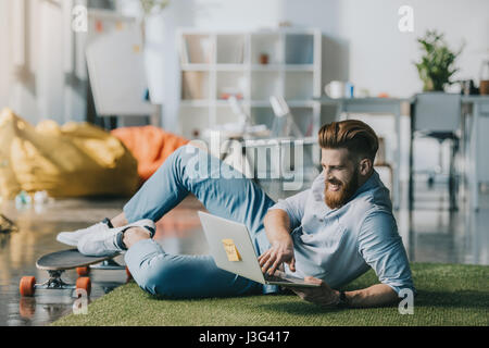 Homme barbu souriant lying on floor and using laptop in creative office Banque D'Images