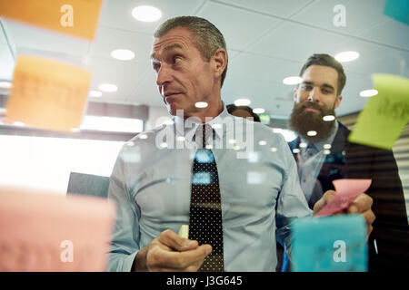 Serious businessman looking at sticky notes sur le verre, le collègue en arrière-plan Banque D'Images