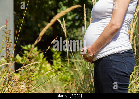 Photo de femme enceinte par le côté, le visage n'est pas visibles. femme est berçant son ventre enceinte, debout à l'extérieur dans un jardin. Banque D'Images