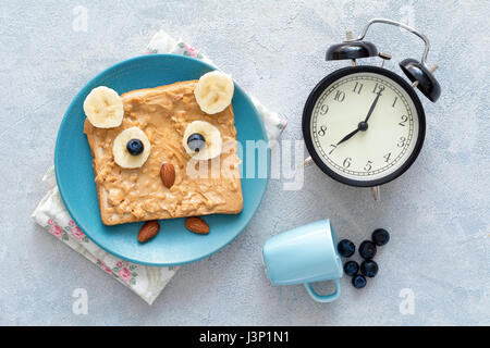 Funny owl toast avec du beurre d'arachide, banane, les bleuets et les amandes sur une plaque bleue, une horloge et petite tasse de bleuets. Vue d'en haut Banque D'Images