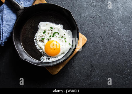 Œuf frit. Vue rapprochée de l'œuf frit à la poêle. Œuf frit épicé et salé avec du persil sur le poêle en fonte et fond noir. Banque D'Images