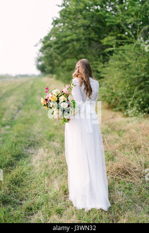 Bouquet de fleurs, les gens, les fêtes , don et arrangement floral concept - belle femme dans une robe blanche à la mode posant avec son dos avec bouquet de fleurs fraîches sur fond d'arbres. Banque D'Images