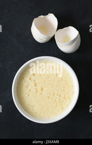 Les oeufs entiers battus (blancs et jaunes ensemble) dans un bol, avec des coquilles d'œufs sur le côté, photographié dans le ciel ardoise avec lumière naturelle Banque D'Images