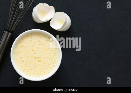 Les oeufs entiers battus (blancs et jaunes ensemble) dans un bol, avec un fouet, et coquilles d'œufs sur le côté, photographié dans le ciel ardoise avec lumière naturelle Banque D'Images