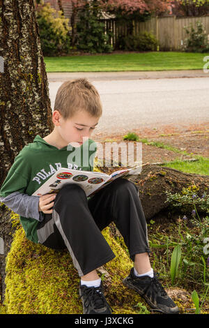 Garçon de neuf ans, la lecture d'un livre à l'ombre d'un arbre en Issaquah, Washington, USA Banque D'Images