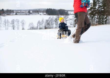 Père Fils tirant le long sur un traîneau, exerçant son jeune garçon dans les bras, dans un paysage couvert de neige, low section Banque D'Images