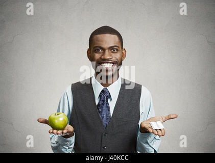 Closeup portrait young man holding exécutif fit Happy apple vert frais dans une main, des pilules, des vitamines dans l'autre, essayant de décider quel choix est le Banque D'Images