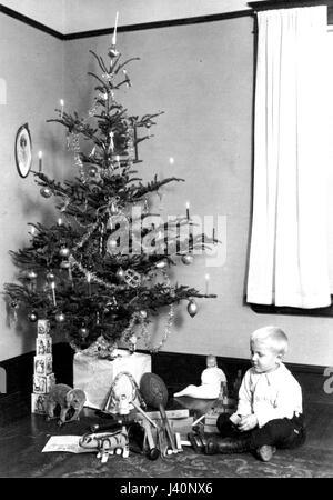 Un jeune garçon assis à côté de l'arbre de Noël familial, qui a éclairé des bougies, c. De nombreux cadeaux sont mis en valeur, y compris un éléphant jouet, des outils de jouet, un football, des blocs pour enfants, un pavillon de jouet, une poupée, etc. Pour voir mes autres images vintage liées à Noël, recherche: Vacances vintage de préstor Banque D'Images