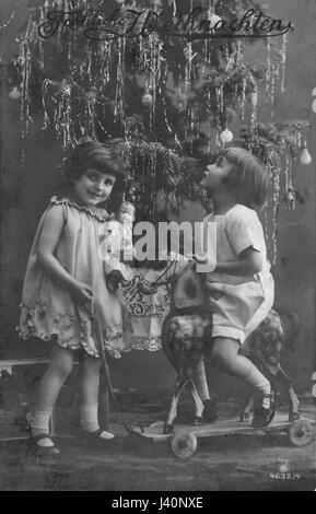 Deux jeunes filles, profitant d'un cheval hobby rideable et d'une fine poupée allemande comme cadeaux de Noël, devant un arbre de Noël avec des ornements et des ornements. 1913 pour voir mes autres images d'époque liées à Noël, recherchez: Prestor vacances d'époque Banque D'Images