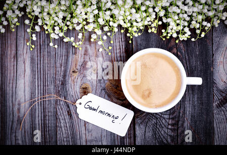 Tasse blanche avec du café sur une surface en bois gris, vue du dessus, à proximité d'un tag avec une inscription bonjour, décoration avec des fleurs de lys de la vallée Banque D'Images