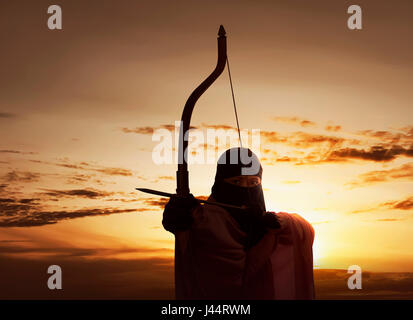 Asian Muslim woman wearing hijab holding bow de tirer une flèche contre fond coucher de soleil Banque D'Images