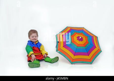 Bébé garçon, vêtu d'un costume coloré plus fluide et des bottes en caoutchouc, est assis à côté d'un parasol et de pleurer. Banque D'Images