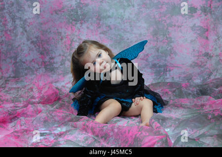 Adorable petite fille est vêtu d'un costume de fée et assis dans une pièce remplie de rose et gris. Ses yeux sont bleu vif et correspondre à ses Ailes et jupe Banque D'Images