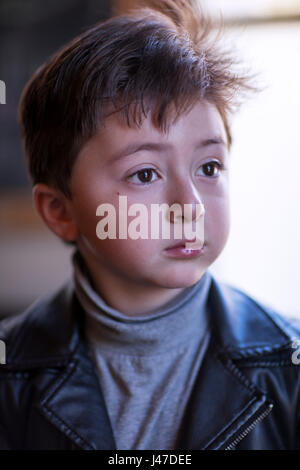 Beau petit garçon aux grands yeux marron et les cheveux bruns épais portant une chemise à col roulé gris et noir veste moto en cuir Banque D'Images