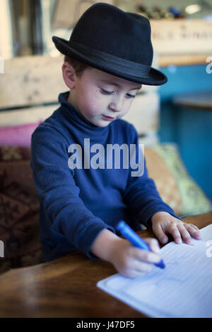 Mignon petit garçon avec les cheveux bruns portant un chapeau fedora noir et un pull à col roulé bleu dans un livre de coloriage avec un crayon crayola bleu Banque D'Images