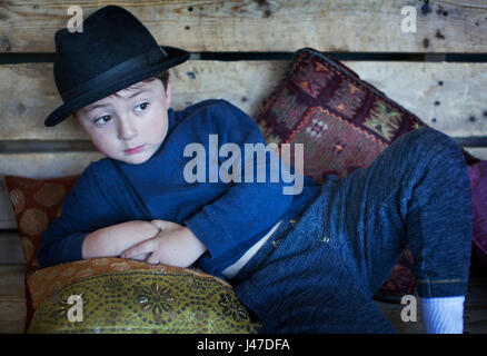 Mignon petit garçon avec les cheveux bruns portant un chapeau fedora noir et un pull à col roulé bleu couchée sur un banc en bois avec des oreillers Banque D'Images