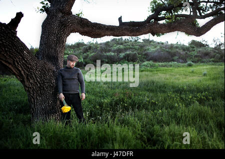 Irlandais en laine pull gris col roulé avec un bouchon jaune brun crieur holding bouquet de fleurs debout dans un champ d'herbe verte avec Oak tree Banque D'Images