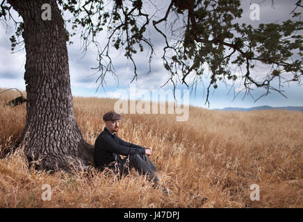 Triste homme solitaire avec une barbiche rouge portant un jean denim noir et chemise de cow-boy chapeau gavroche marron assis sous un grand chêne dans une herbe jaune Banque D'Images