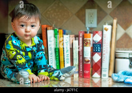 Mignon et happy baby boy wearing pyjamas colorés assis dans la cuisine en face de nombreux livres de cuisine Banque D'Images