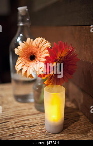 Daisy orange et rouge fleurs en pot de verre ballon entre vintage bouteille d'eau et petite bougie sur la table en bois Banque D'Images