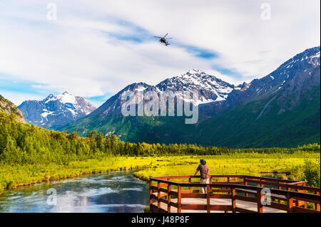 Un homme est debout sur la promenade du centre naturel d'Eagle River tandis Qu'Un hélicoptère Black Hawk survole Head dans le parc national de Chugach à Southcentra... Banque D'Images