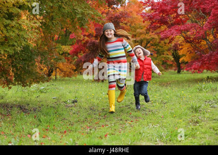 Un jeune garçon et une jeune fille portant des bottes en caoutchouc et des vêtements colorés traversent Un champ tenant les mains avec des arbres dans les couleurs vives de l'automne dans le Backgr... Banque D'Images