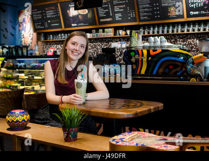 BRASOV, Roumanie - 26 Avril 2017 : Young blonde woman smiling s'asseoir à une table en verre café shop local frappe avec mousse Banque D'Images