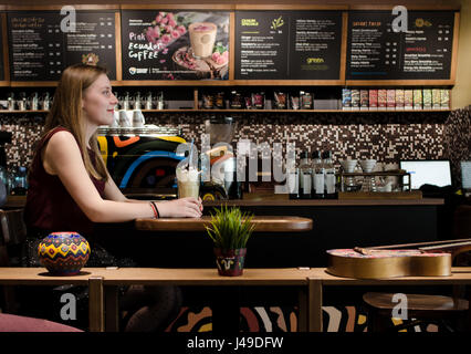 BRASOV, Roumanie - 26 Avril 2017 : Côté de young blonde woman smiling s'asseoir à une table en verre café shop local frappe avec mousse Banque D'Images