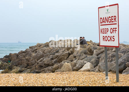 Danger, restez à l'extérieur du panneau avec un couple assis sur des rochers embrassant à Weymouth, Dorset UK en mai Banque D'Images