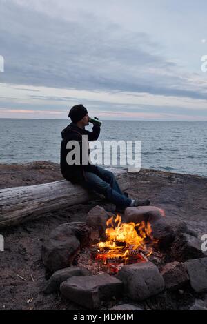 Un homme avec une bière assis près d'un feu de camp au lac Supérieur dans le Minnesota, USA. Banque D'Images