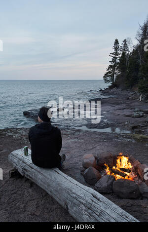 Un homme avec une bière assis près d'un feu de camp au lac Supérieur dans le Minnesota, USA. Banque D'Images