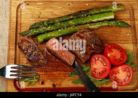 Couper tranches de bifteck cuit moyen juteux servi sur planche de bois avec salade de roquette vert frais, asperges et tomates cerises rouges, fourchette et Banque D'Images