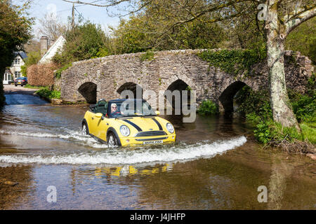 Une femme au volant d'un cabriolet Mini Cooper à travers la Ford à côté du pont médiéval à cheval traversant le fleuve en Haddeo Exmoor. Banque D'Images