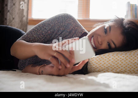 Mixed Race woman laying on bed texting on cell phone Banque D'Images