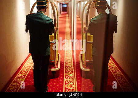 Vue arrière avec miroir portrait reflet de groom poussant chariot à bagages dans couloir de l'hôtel, offrant des chambres à sacs Banque D'Images