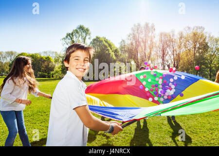 Portrait of happy boy holding parachute arc-en-ciel plein de boules colorées et de jouer avec ses amis dans le parc Banque D'Images