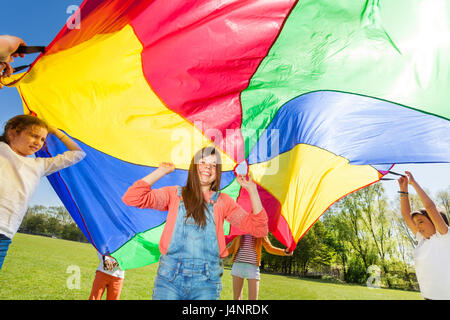 Groupe d'amis heureux de jouer, de se cacher sous parachute fille coloré dans la canopée parc d'été Banque D'Images