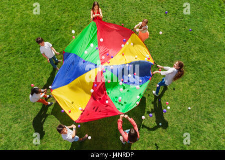 Vue d'en haut photo de sept enfants debout dans un cercle et arc-en-ciel ondulant parachute afin de jeter les boules colorées dans l'air Banque D'Images
