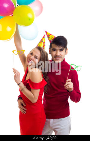 Jeune femme élégante avec de longs cheveux châtains en robe rouge et les jeunes bel homme en chemise rouge avec la célébration de chapeaux détenant plusieurs ballons colorés et c Banque D'Images