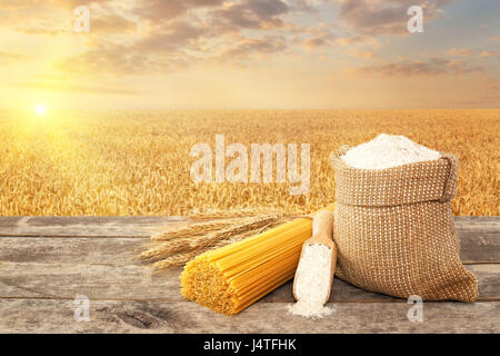 Pâtes alimentaires non cuites à partir de blé dur, farine de grains entiers dans le sac et scoop sur table avec champ de céréales mûrs sur l'arrière-plan. Champ de blé doré sur le coucher du soleil. Ph Banque D'Images