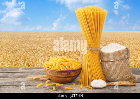 Pâtes alimentaires non cuites, en forme de spirale et spaghetti, de farine de farine en sachet et la cuillère sur la table avec champ de céréales sur l'arrière-plan. Champ de blé doré w Banque D'Images