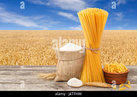 Macaroni cru, en forme de spirale et spaghetti, de farine de farine dans le sac sur table avec champ de céréales mûrs sur l'arrière-plan. Champ de blé doré avec ciel bleu Banque D'Images