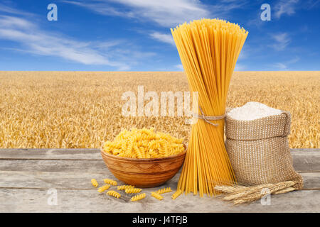 Pâtes alimentaires non cuites, en forme de spirale et spaghetti, de farine de farine dans le sac sur table avec champ de céréales mûrs sur l'arrière-plan. Champ de blé doré avec blue s Banque D'Images