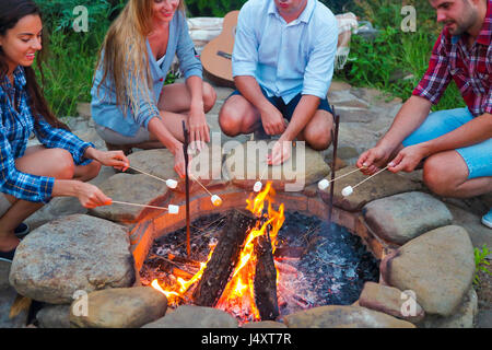 Compagnie des jeunes gens tenant des bâtons avec des guimauves au-dessus du feu de camp en soirée. Maison de vacances et d'amitié concept Banque D'Images