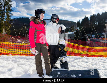 Jeune couple heureux en combinaisons de ski, casques et lunettes de ski debout avec les planches près de la clôture dans une station de ski en hiver Banque D'Images