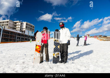 Jeune couple en combinaisons de ski, casques et lunettes de ski debout avec les planches dans une station de ski en hiver Banque D'Images